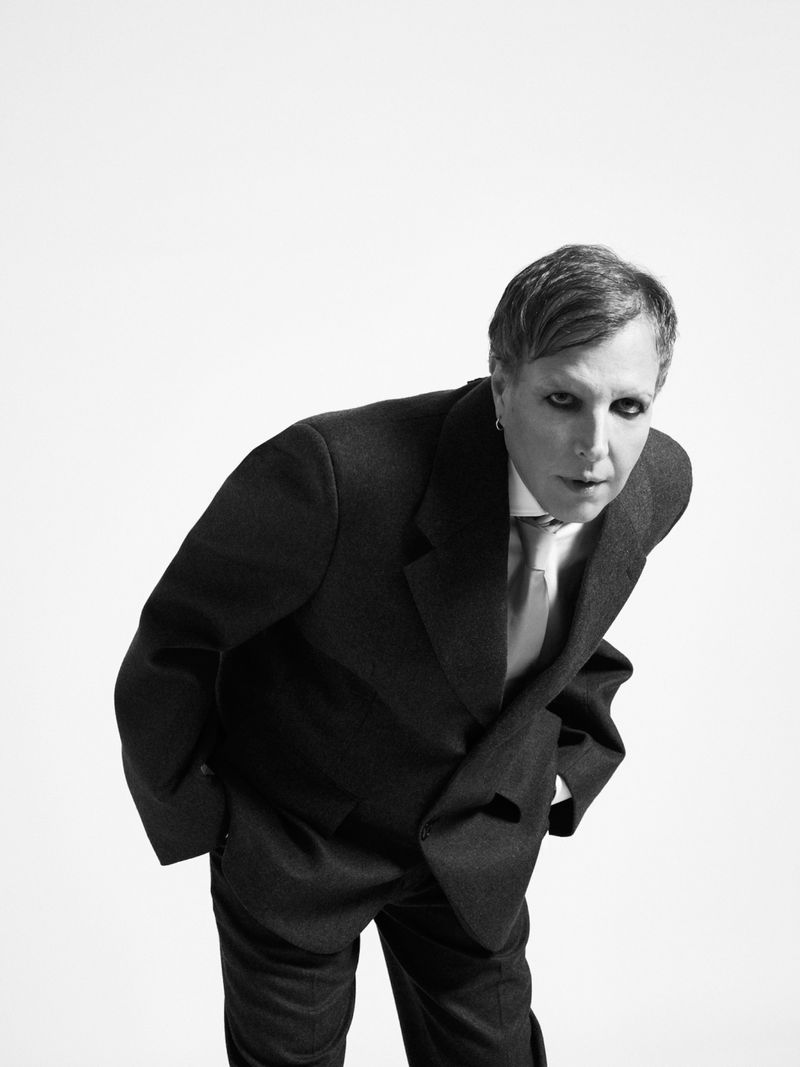Person in a dark tailored suit and tie leaning forward against a seamless white background