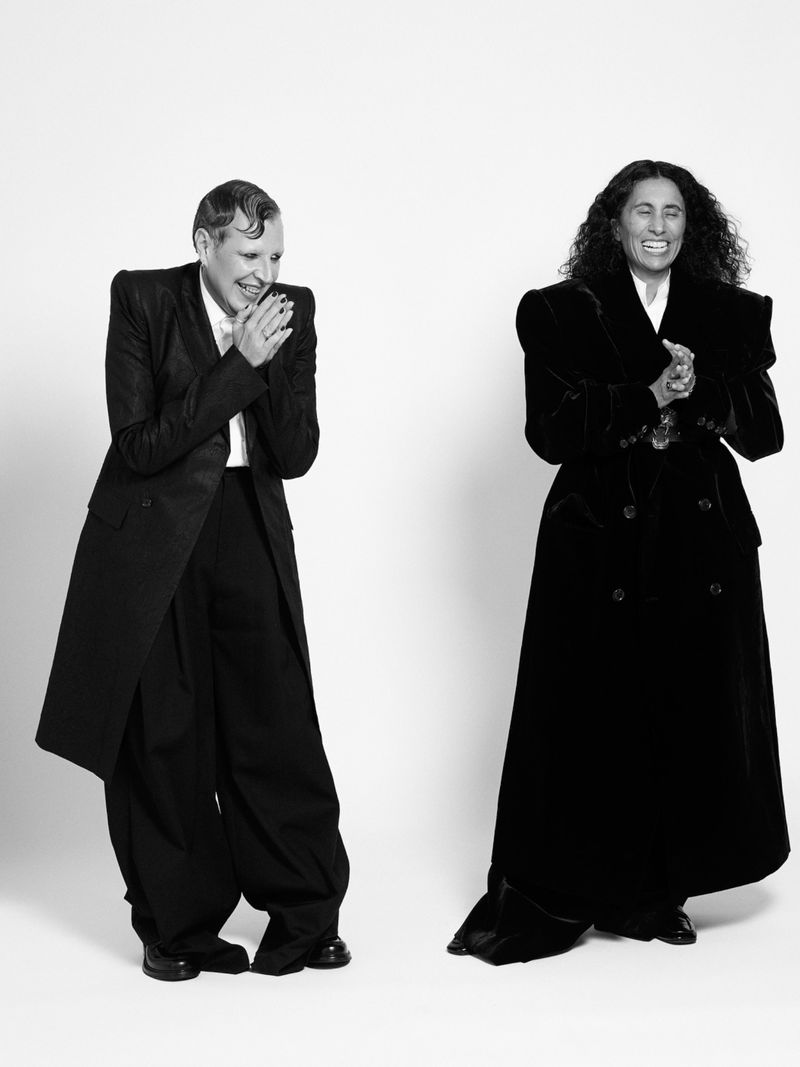 Two stylish adults in black coats pose in a black-and-white studio shot against a plain white backdrop, radiating high fashion.