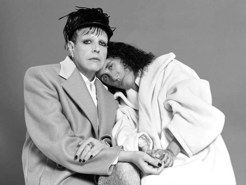 Black and white portrait of two people, one with a hat and tailored jacket, the other in a white robe, holding hands against a gray background.