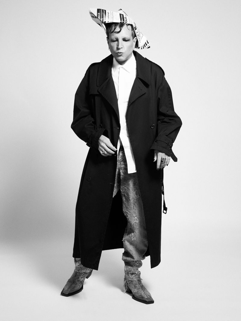 A person in a long black coat and silver pants stands in front of a white background wearing a newspaper hat in a studio setting.