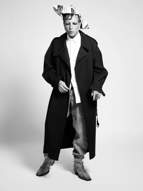 A person in a long black coat and silver pants stands in front of a white background wearing a newspaper hat in a studio setting.