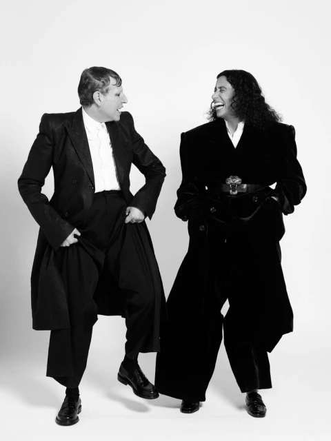 Two adults in elegant black outfits laugh together in a black-and-white studio shot, showcasing retro-inspired fashion and joyful expression.