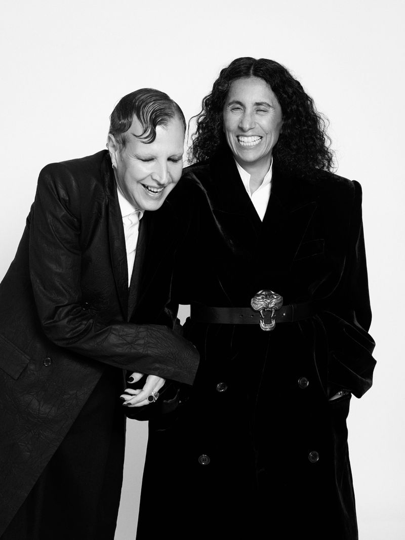 Black-and-white portrait of two adult females smiling in a photo studio. One woman wears a blazer, the other a belted trench coat.