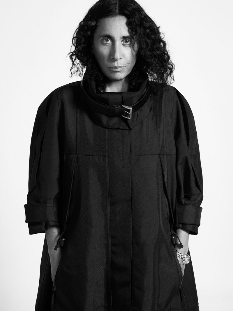 Monochromatic portrait of a dark-haired woman with curly hair wearing a black coat against a bright white studio backdrop, fashion.