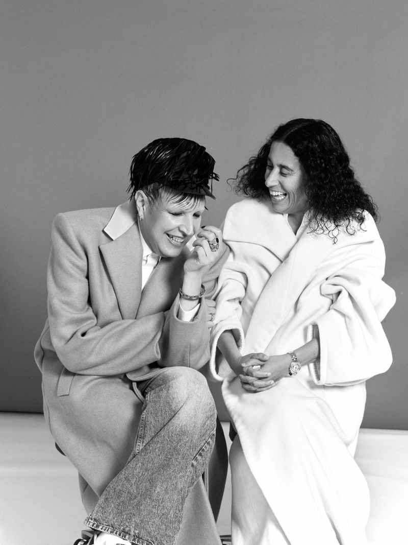 Two people seated against a grey backdrop, one wearing a tailored coat and jeans, the other in a light robe, laughing.