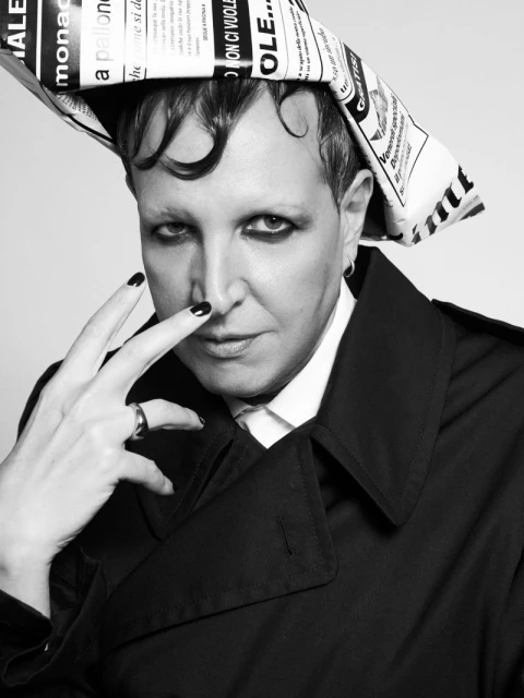 Adult with dark hair, wearing a dark overcoat and newsprint hat, poses indoors against a white background in a black and white portrait.