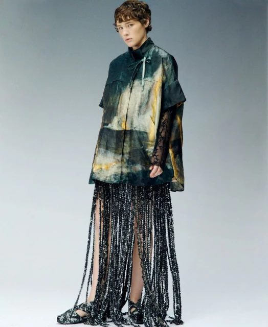 Full-length studio shot of a slender person with short hair wearing an abstract patterned top and fringed skirt with matching shoes.