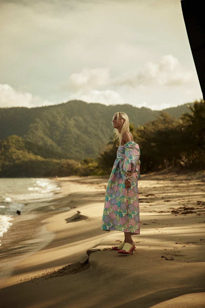 A woman in a floral dress stands on a sandy beach with mountains in the background, creating a scenic outdoor fashion shot.
