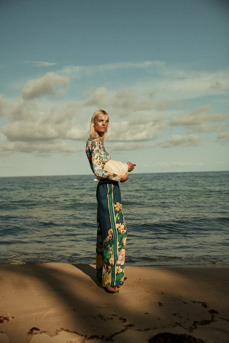 Model standing on a sandy beach wearing patterned floral long-sleeve top and matching trousers against the ocean