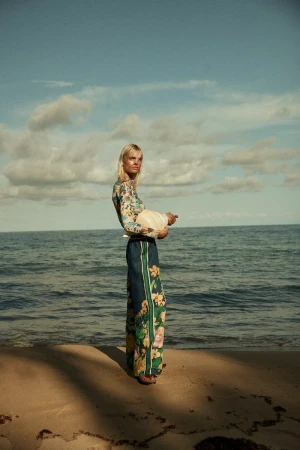 Model standing on a sandy beach wearing patterned floral long-sleeve top and matching trousers against the ocean