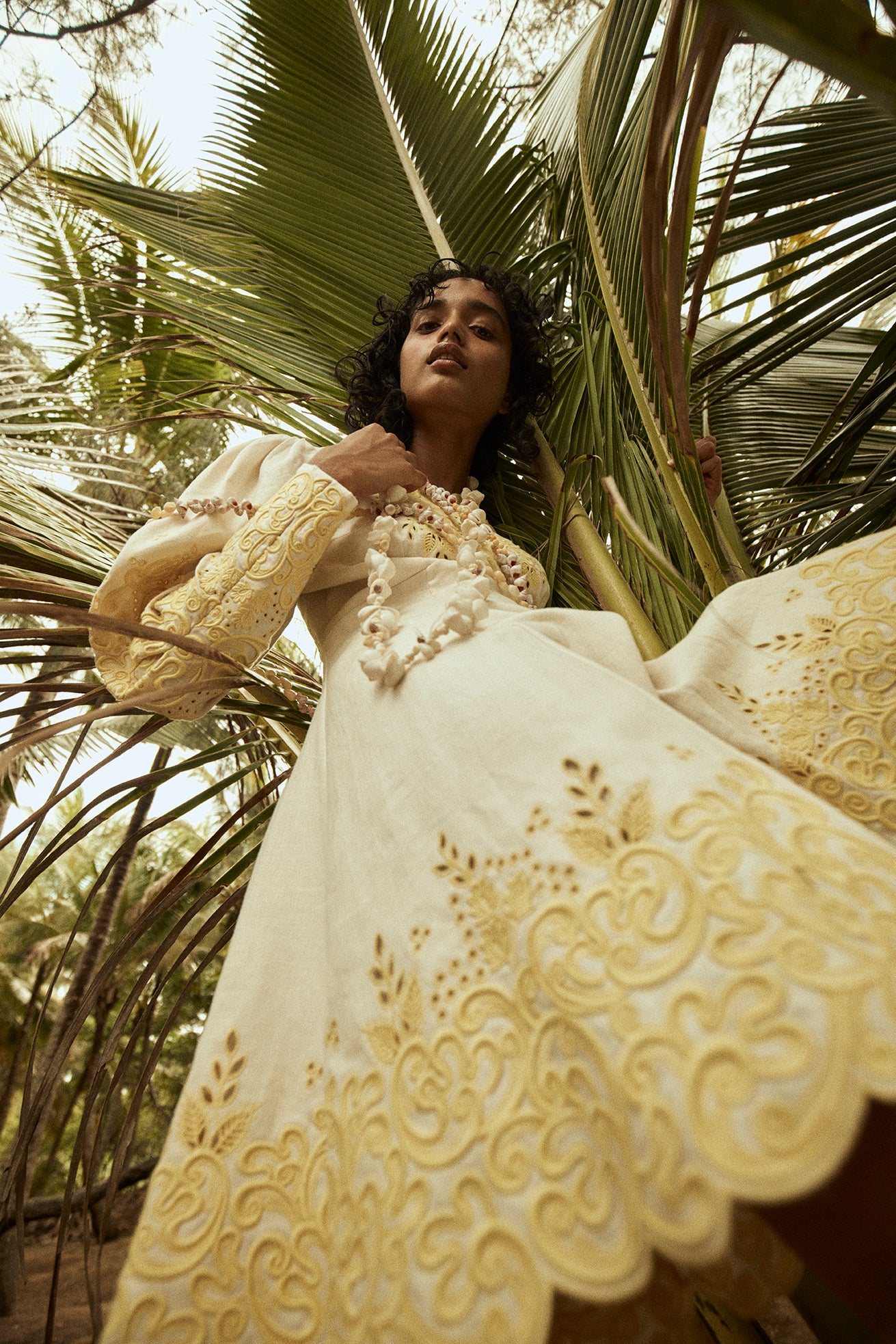 Woman poses in a cream embroidered dress surrounded by palm fronds in an outdoor setting, low angle view.