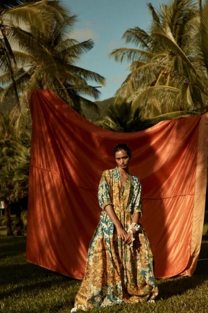 Woman in flowing floral dress poses in front of a copper-colored cloth backdrop, with palm trees in the background and green grass.