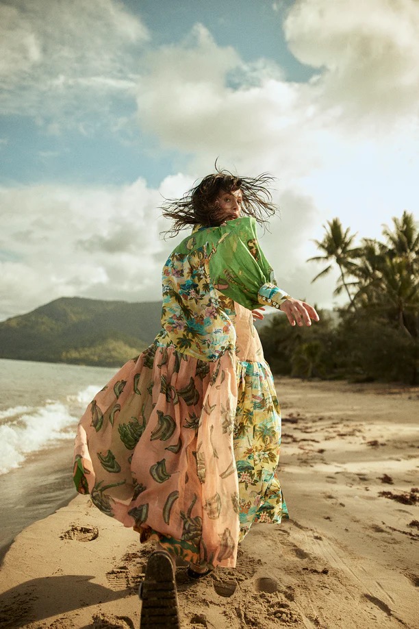 Woman in a tropical print dress walks on a sandy beach with palm trees and mountains in the background under a partly cloudy sky.