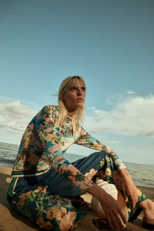 Model with blonde hair wearing a floral print long-sleeve top and blue pants seated on a sandy beach against an ocean and sky backdrop.