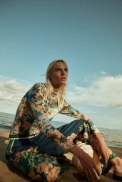 Model with blonde hair wearing a floral print long-sleeve top and blue pants seated on a sandy beach against an ocean and sky backdrop.