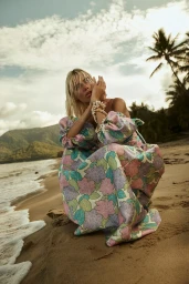 Model in a floral print dress crouching on a sandy beach with palm trees under an overcast sky.