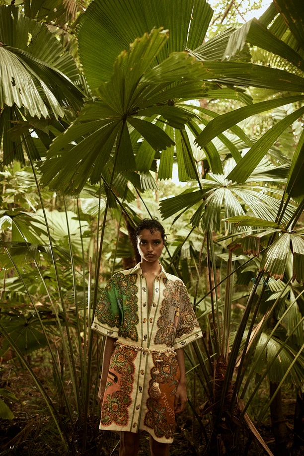 Fashionable woman in paisley dress poses in lush jungle setting surrounded by tropical palm leaves and dense green foliage.