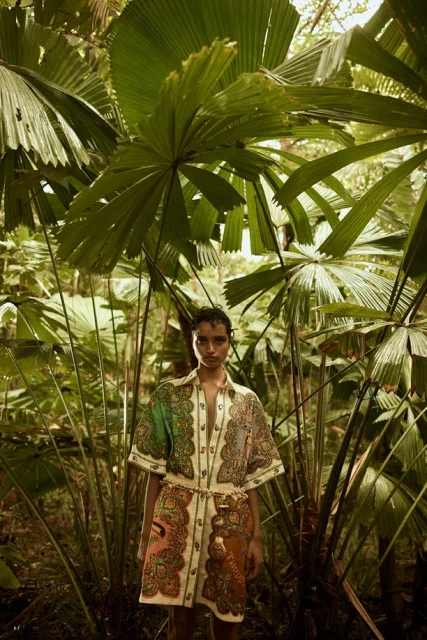 Fashionable woman in paisley dress poses in lush jungle setting surrounded by tropical palm leaves and dense green foliage.