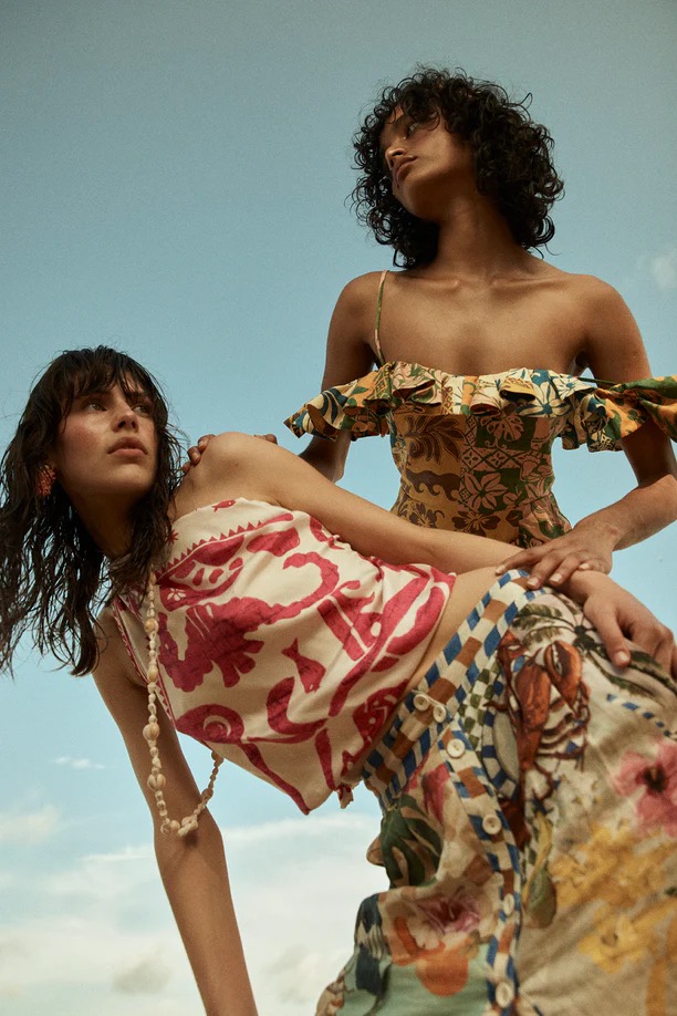 Two adult females pose outdoors in colorful floral-patterned dresses against a blue sky, showcasing fashion and style in a medium shot.