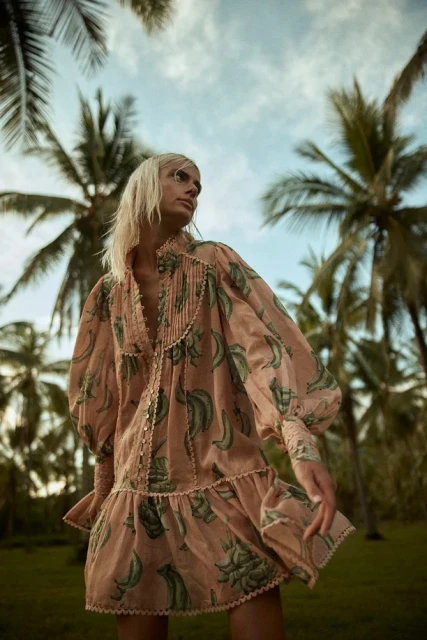 A fair-skinned model poses in a banana-printed mini-dress in a tropical environment surrounded by palm trees and a blue sky.