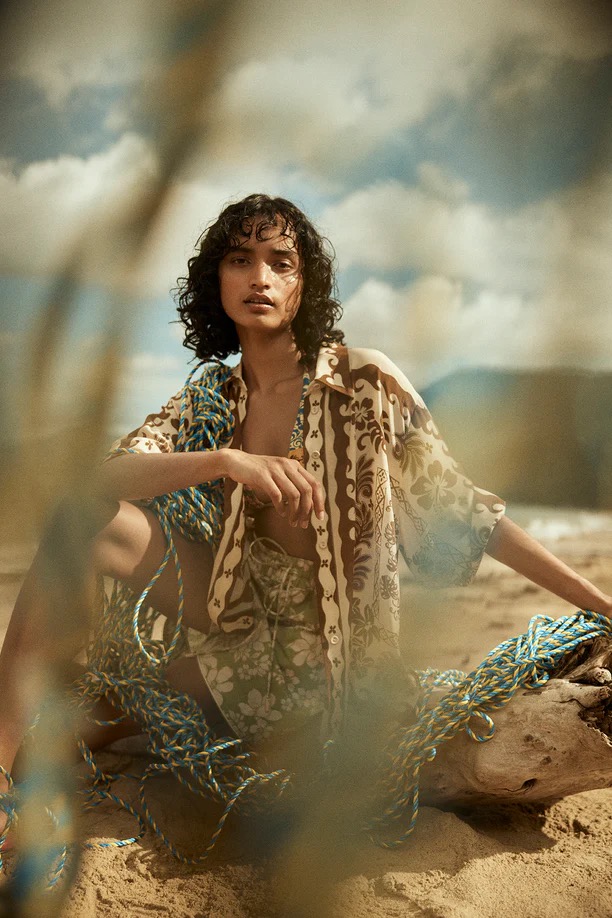 Woman in patterned shirt and shorts sits on the beach with a coil of rope and a blue sky with clouds in the background. 