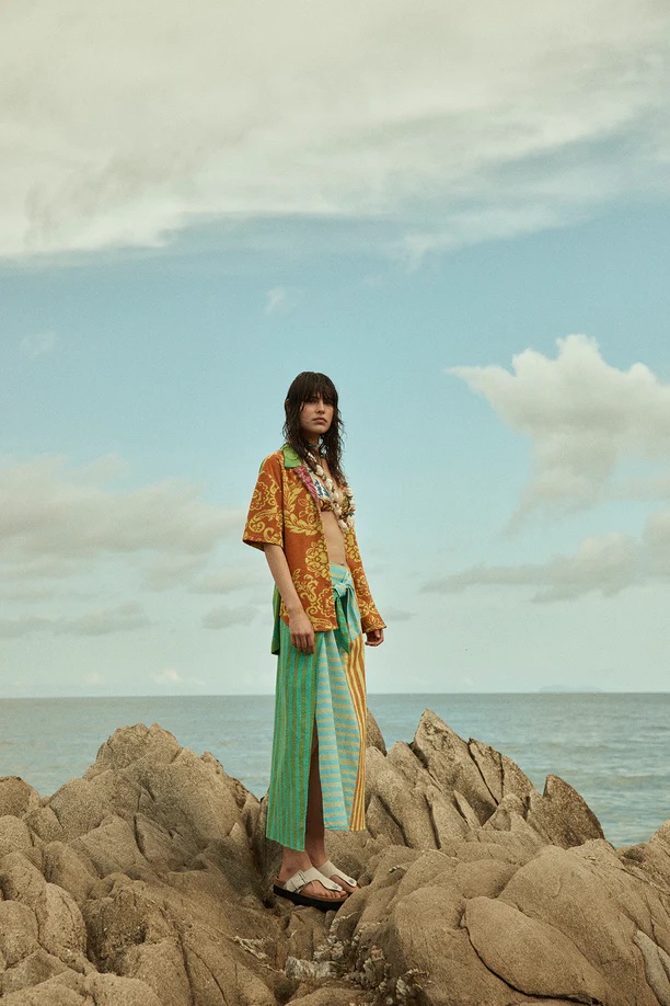 A woman poses on rocks by the sea, wearing a patterned shirt, sarong, and sandals under a cloudy sky, showcasing fashion and nature.