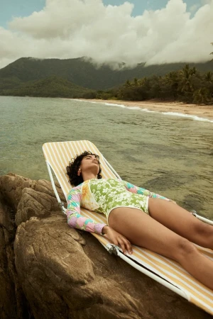 Woman in floral swimsuit reclines on striped lounge chair near ocean with mountains in background, enjoying a sunny day.