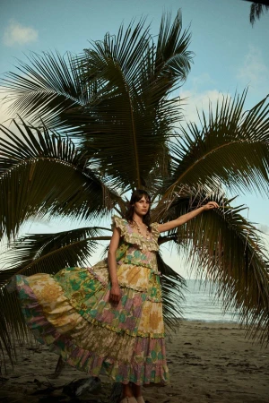 A woman wearing a tiered floral dress standing beneath a palm tree on a sandy beach by the ocean.