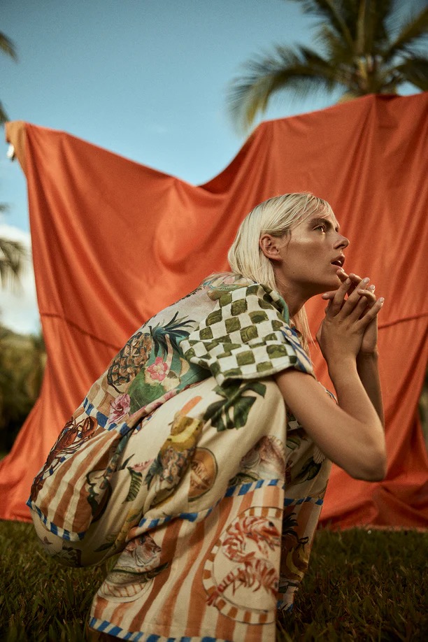 Woman in printed shirt and pants crouching in grassy outdoor area in front of orange fabric, set against palm trees and blue sky.