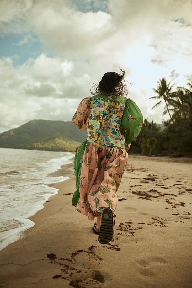 A woman in a colorful floral dress walks along a sandy beach with palm trees, distant mountains, and a cloudy sky in the background.