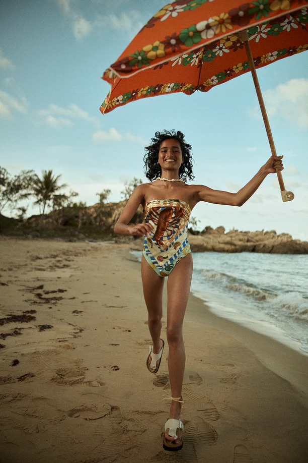 Smiling woman in swimsuit runs on sandy beach with floral umbrella on sunny day; trees, rocks, and sky in background.
