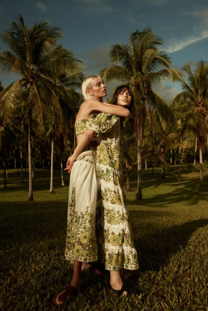 Two adult women pose in floral print matching outfits in a grassy tropical location with palm trees under a partially cloudy blue sky.