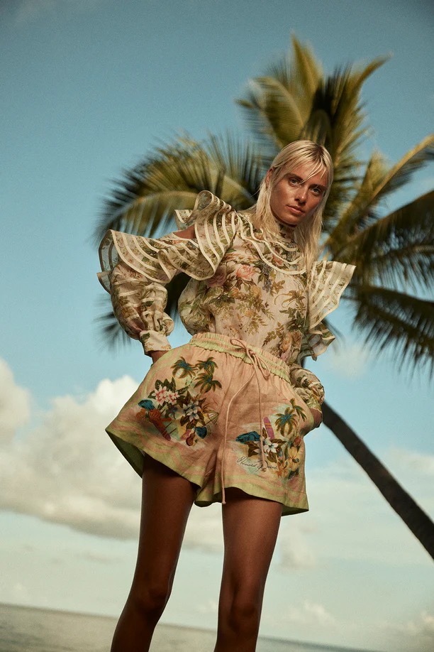 Fashion shoot featuring a blonde woman in tropical print blouse and shorts, posing outdoors against a backdrop of sky, ocean, and palm tree.
