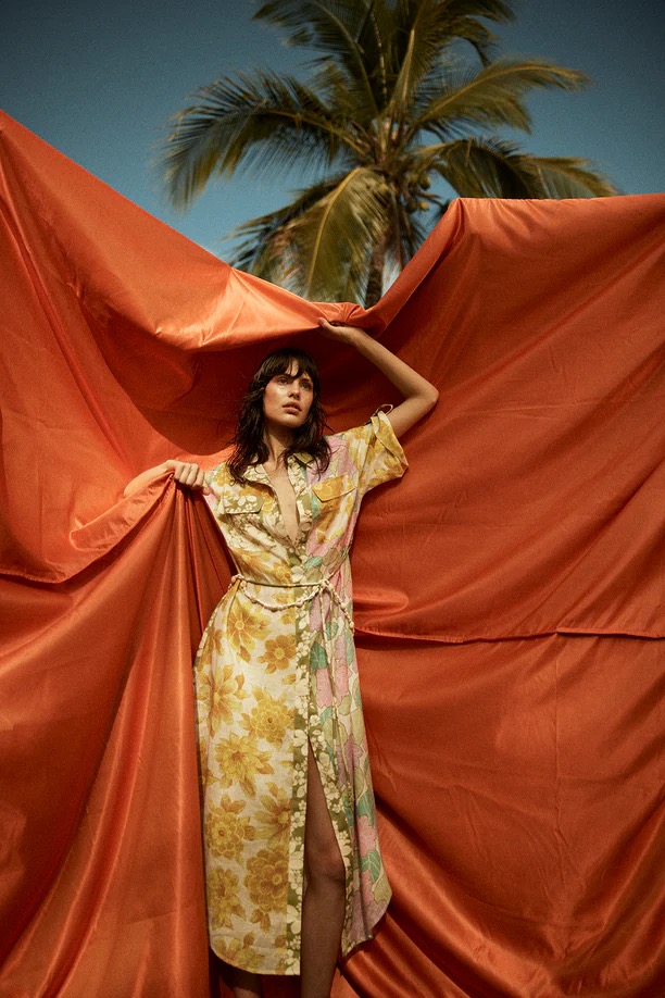 A brunette model in a floral dress poses with an orange sheet in front of a palm tree under a bright blue sky in an outdoor setting.