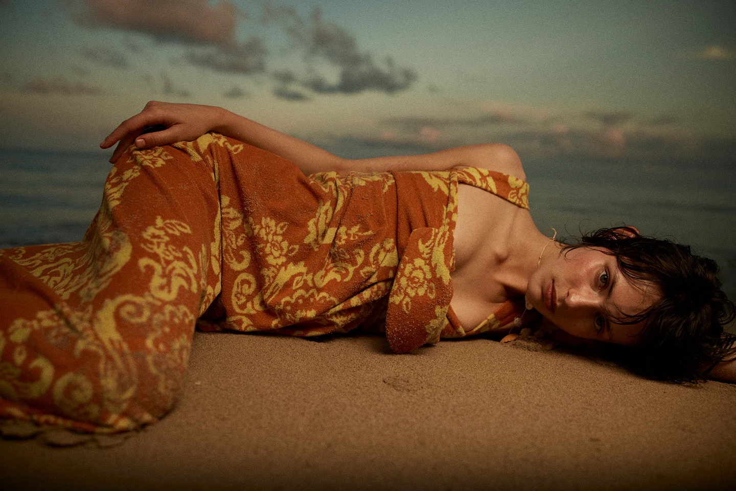 Young woman wrapped in an orange patterned towel, lies on a sandy beach with a soft sunset sky and ocean backdrop, creating a serene image.