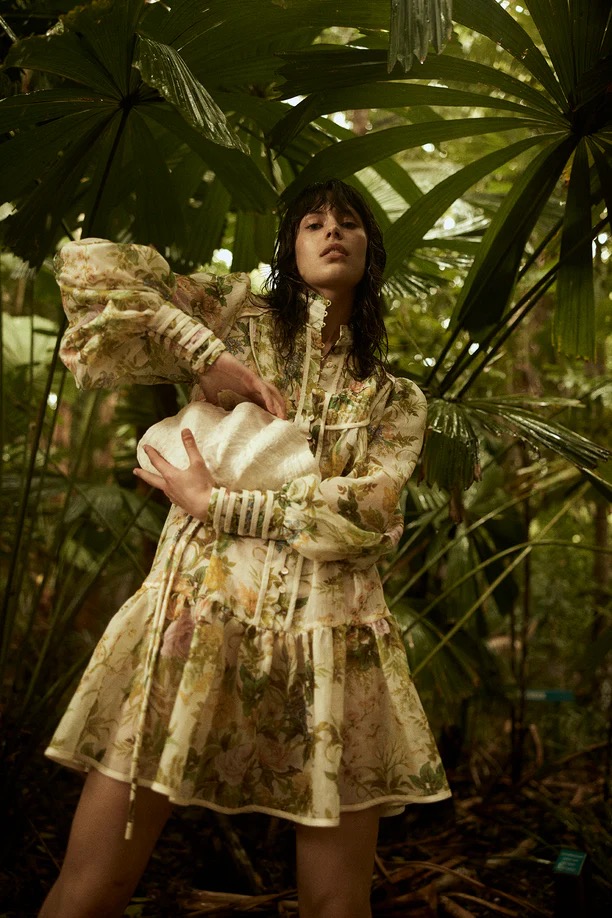 Woman in floral dress holds a white bag in a tropical forest setting. Fashion editorial shot. Exotic plants surround.