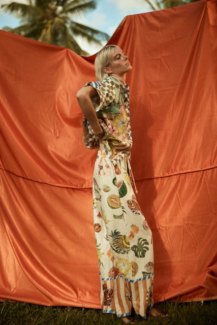 Adult fashion model poses in an outdoor setting wearing a patterned matching shirt and pants in front of a draped orange fabric.
