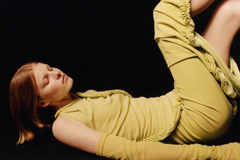 Woman in a chartreuse outfit poses against a dark backdrop. A close-up studio shot captures a fashion pose with soft lighting.