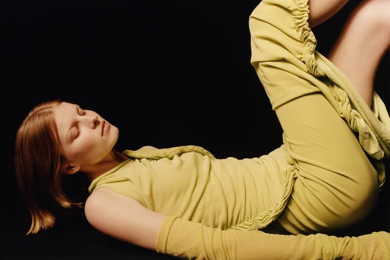 Woman in a chartreuse outfit poses against a dark backdrop. A close-up studio shot captures a fashion pose with soft lighting.
