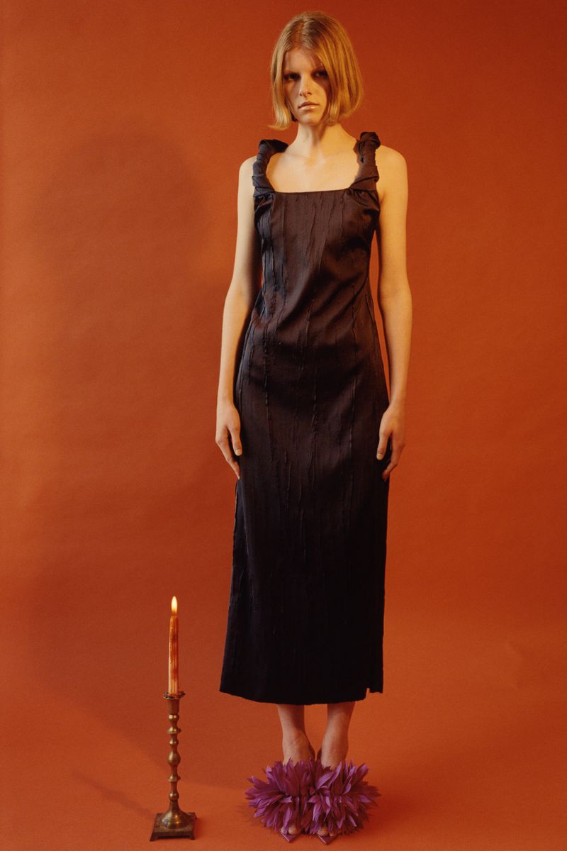 Full shot of an adult female fashion model posing in a dark dress, with a lit candle and flower-like shoes against an orange backdrop.