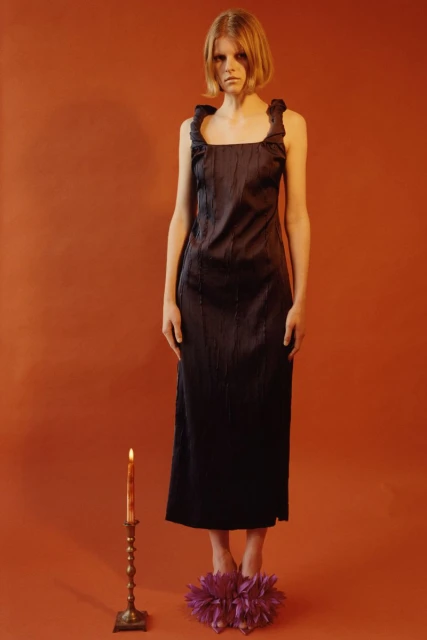 Full shot of an adult female fashion model posing in a dark dress, with a lit candle and flower-like shoes against an orange backdrop.