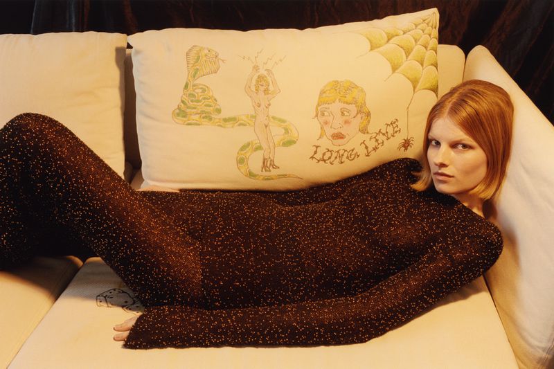 A woman with red hair is lying on a white couch next to a decorative pillow with illustrations in a dimly lit indoor setting.