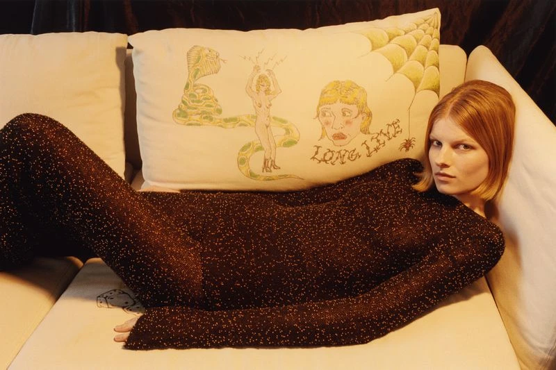 A woman with red hair is lying on a white couch next to a decorative pillow with illustrations in a dimly lit indoor setting.