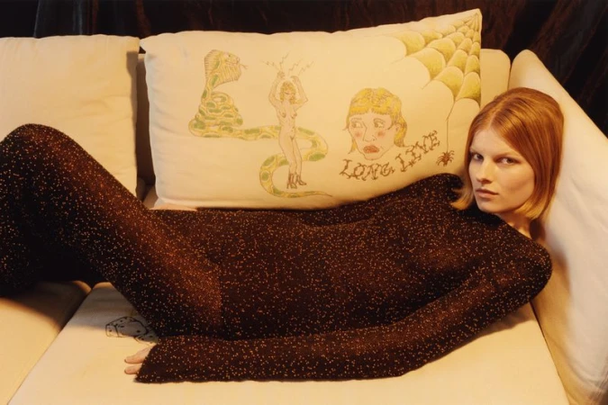 A woman with red hair is lying on a white couch next to a decorative pillow with illustrations in a dimly lit indoor setting.
