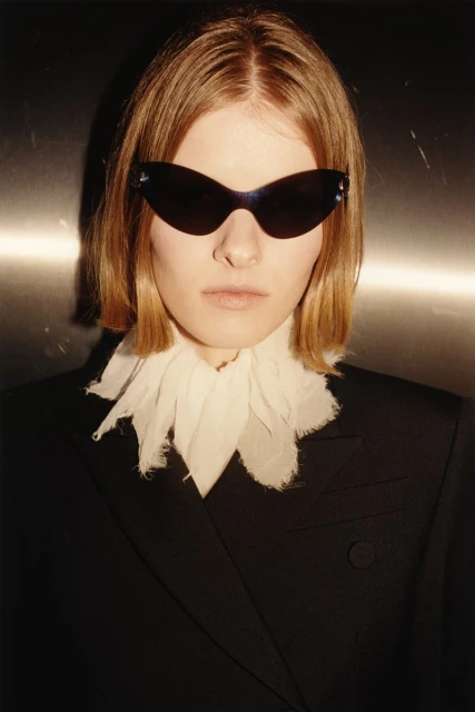 A person with blonde hair wearing a black blazer, white ruffled collar, and sunglasses, looking directly at the camera in an indoor setting.