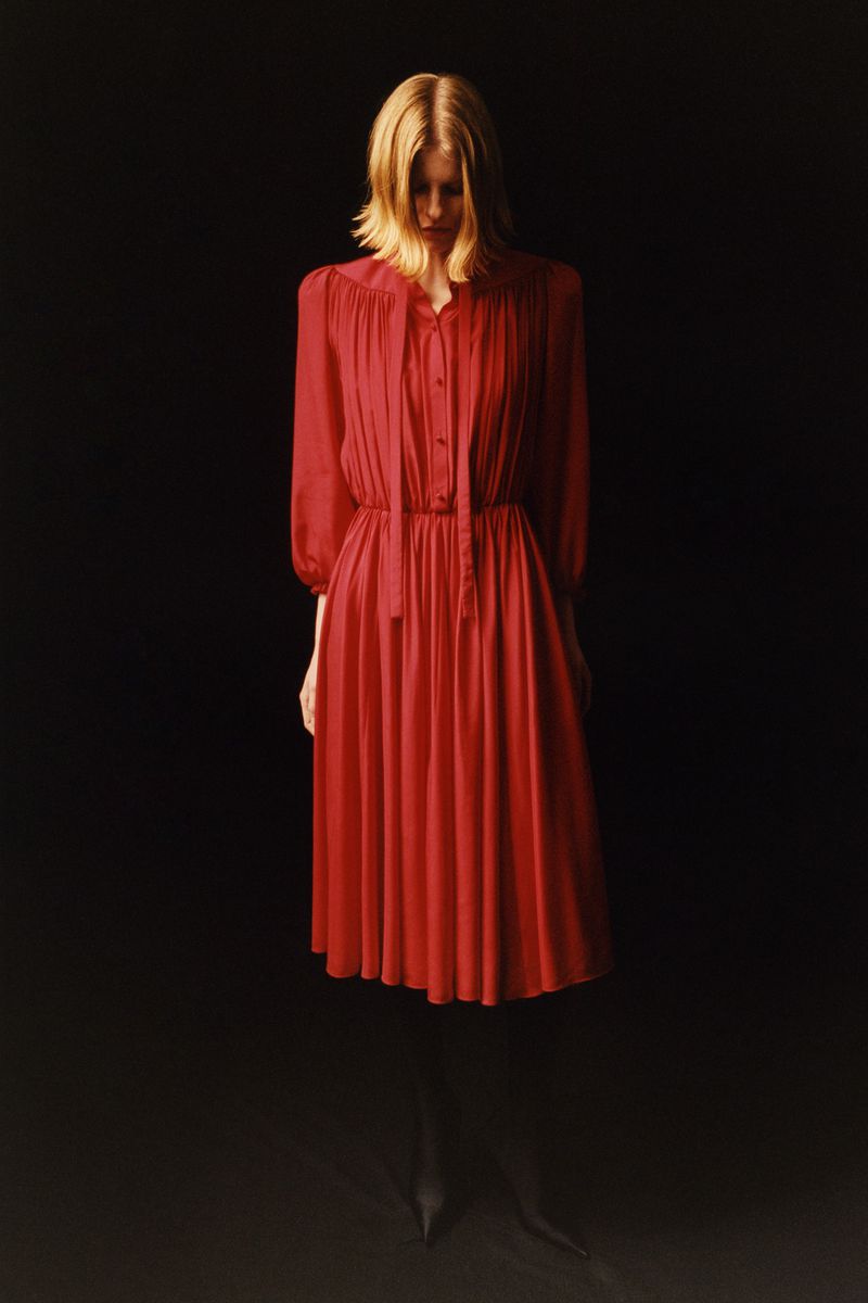 Model standing against a dark background wearing a long-sleeved pleated red dress with a button-up bodice.
