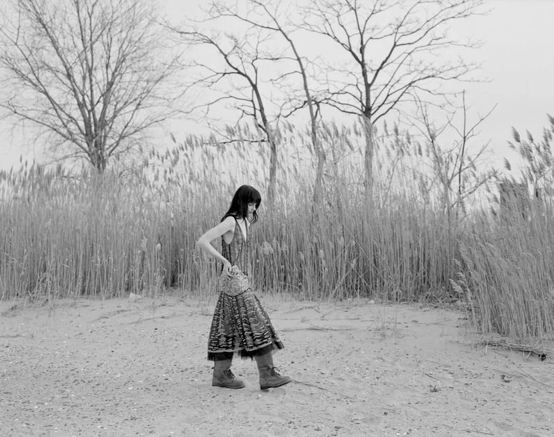 Monochrome outdoor shot of a stylish adult woman wearing patterned dress and boots, standing on sand, with tall grass and trees.