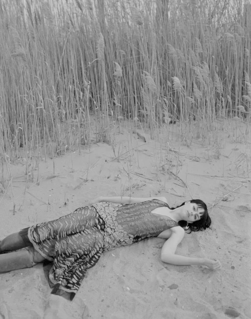 Black and white photograph of a woman in a patterned dress lying on a sandy beach with tall reeds in the background.