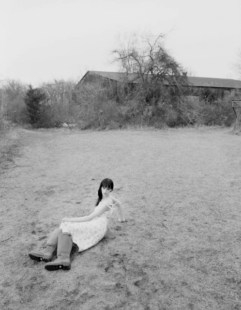 Woman in floral dress and boots reclines in grassy field near barn under overcast sky in black-and-white outdoor fashion shot.