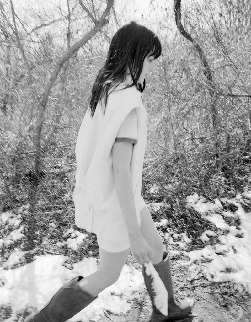 Young woman walks in a snowy forest wearing a light-colored coat, dress, and brown boots, surrounded by bare trees and white snow.